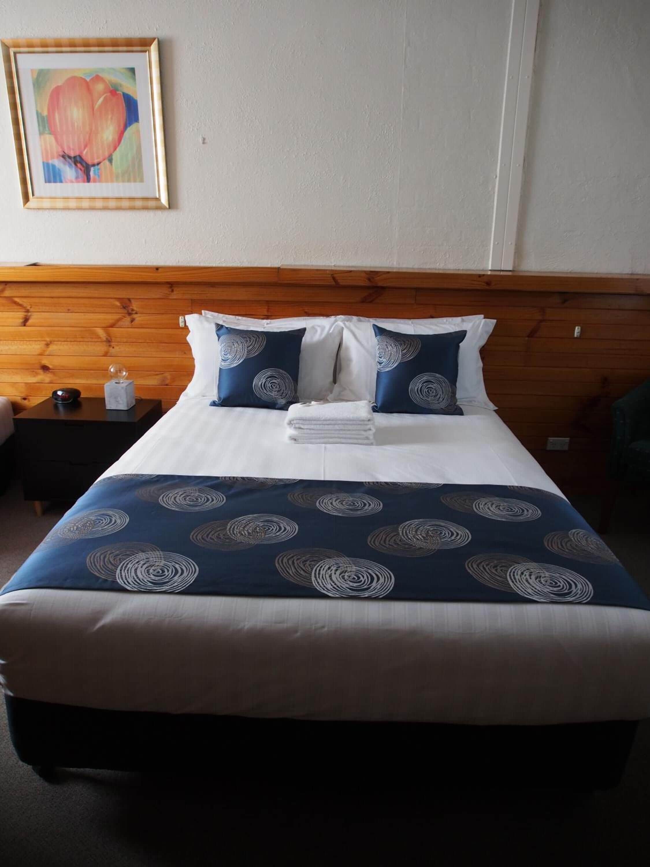 Ein ordentlich bezogenes Bett mit einer blau-weißen Bettdecke, zwei Kissen mit einem kreisförmigen Muster und ein gerahmtes Bild an der Wand.