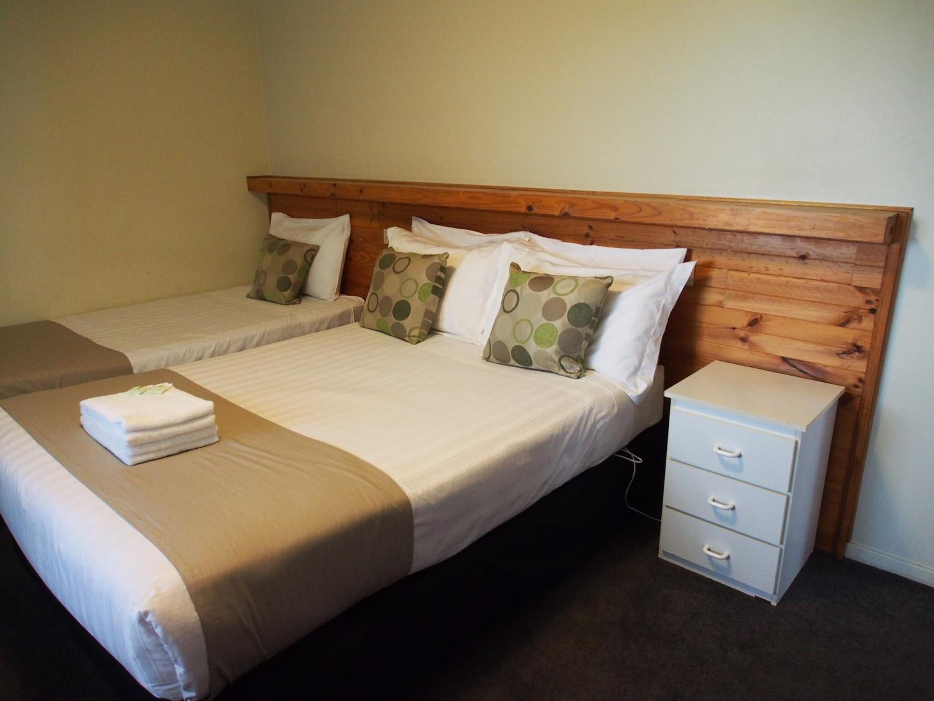 A hotel room with two single beds, a wooden headboard, a small bedside table with drawers, and a neatly made bed with a folded towel.