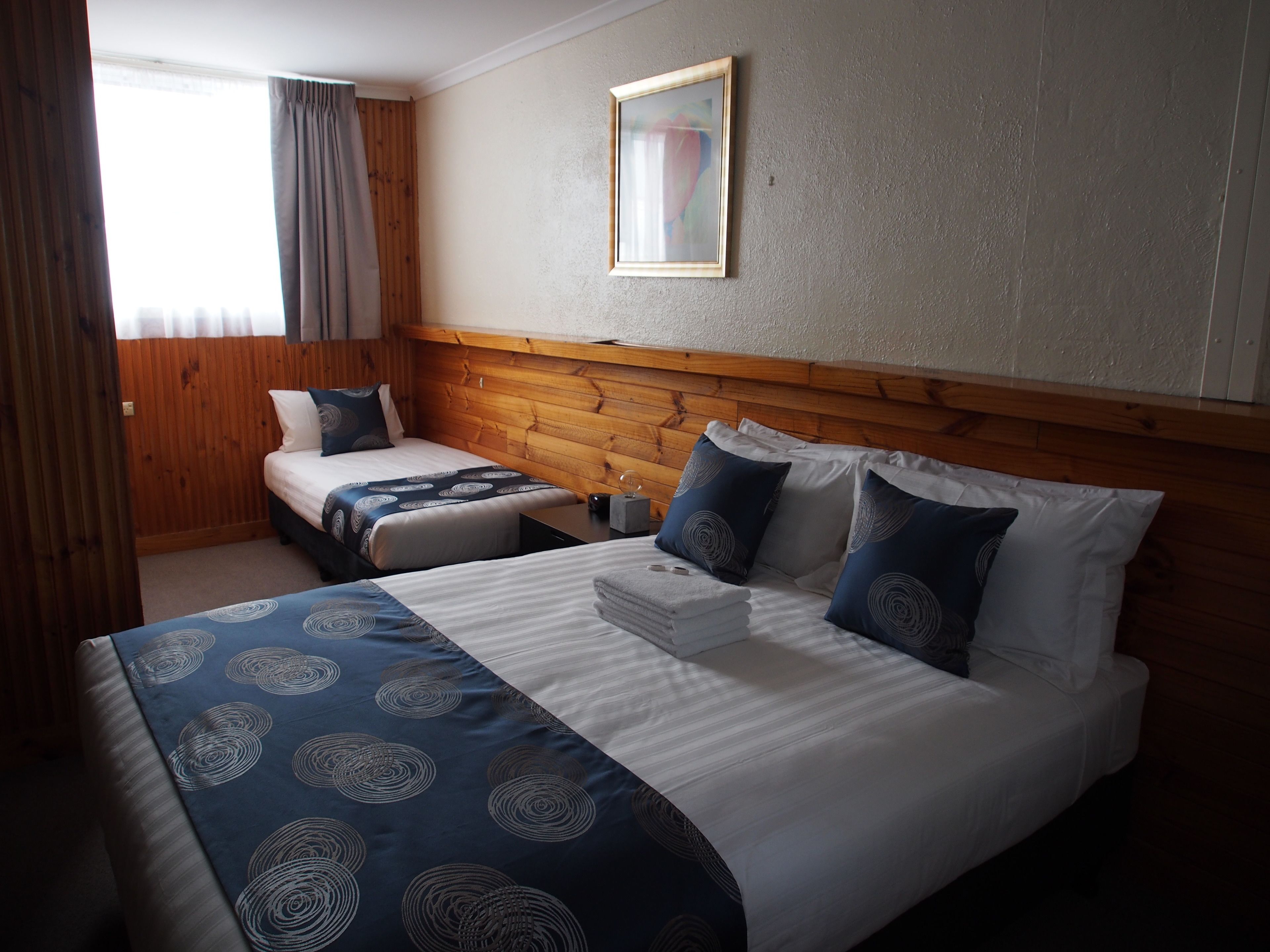 Een hotelkamer met twee bedden, een houten hoofdeinde, een schilderij aan de muur en een raam met gordijnen.