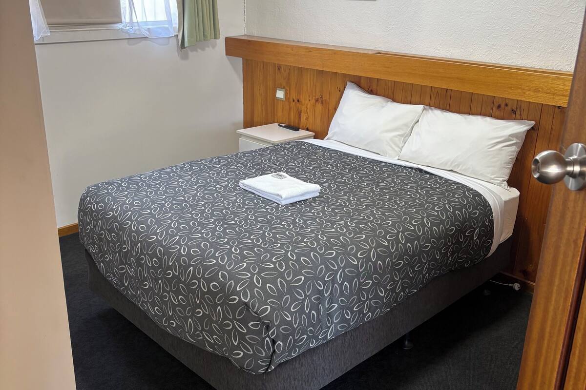 A neatly made bed with a patterned bedspread, a bedside table with a remote control, and a framed picture on the wall.