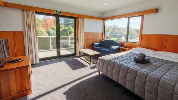 A hotel room with a bed, a desk with a TV and remote, a chair, a small table, and a balcony with a view.