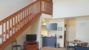 A living room with a wooden staircase, a TV on a wooden stand, a kitchen area with cabinets, a dining table with chairs, and a blue sofa.