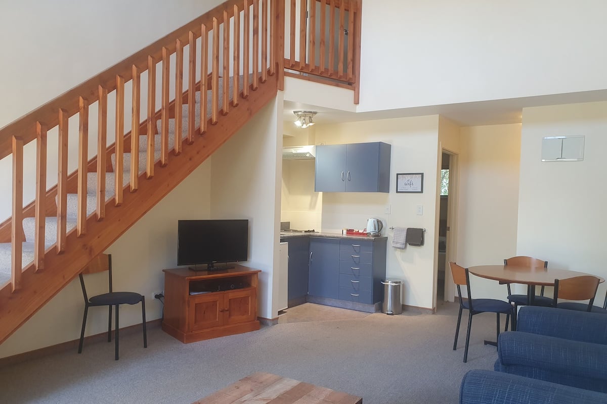A living room with a wooden staircase, a TV on a wooden stand, a kitchen area with cabinets, a dining table with chairs, and a blue sofa.