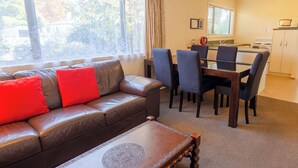 A living room with a brown leather sofa, red cushions, a glass coffee table, and a dining area with a wooden table and chairs.