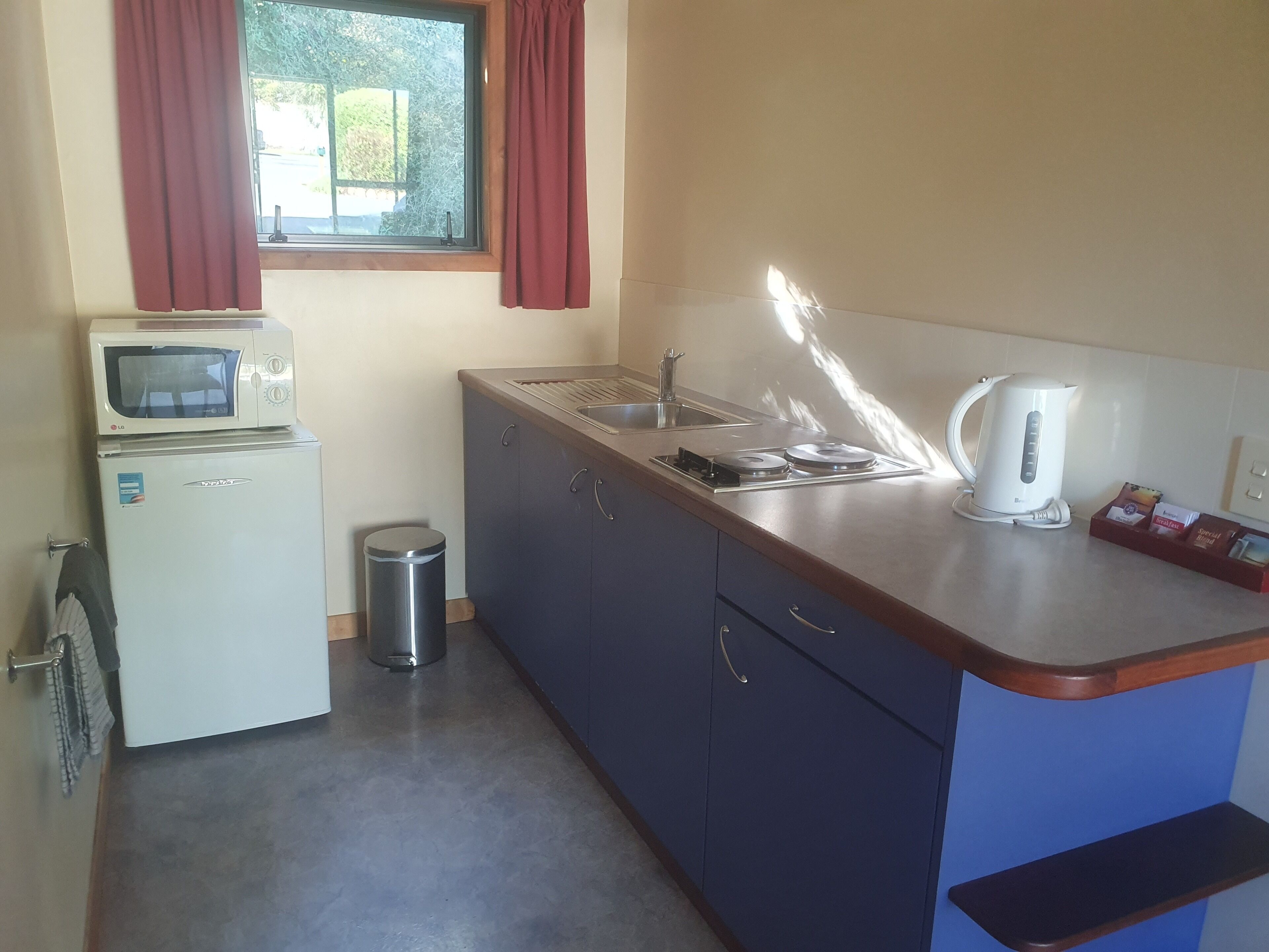 A compact kitchen with a microwave, sink, kettle, and a small trash bin.