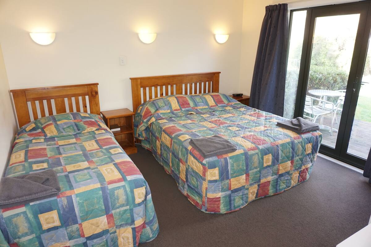 A room with two beds, each with a colorful quilt, a wooden headboard, bedside tables, and a view through a glass door to an outdoor area.