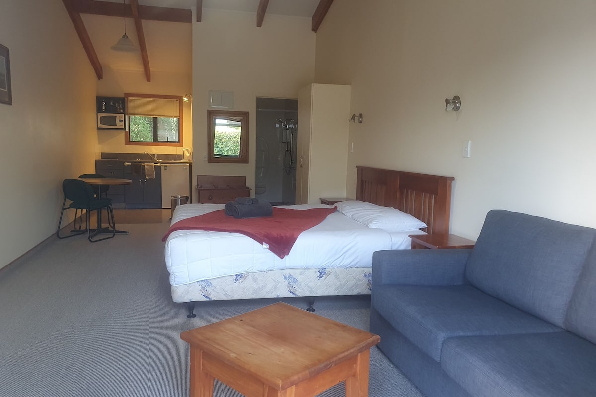 A bedroom with a bed, a wooden nightstand, a blue sofa, a wooden table, and a kitchenette in the background.