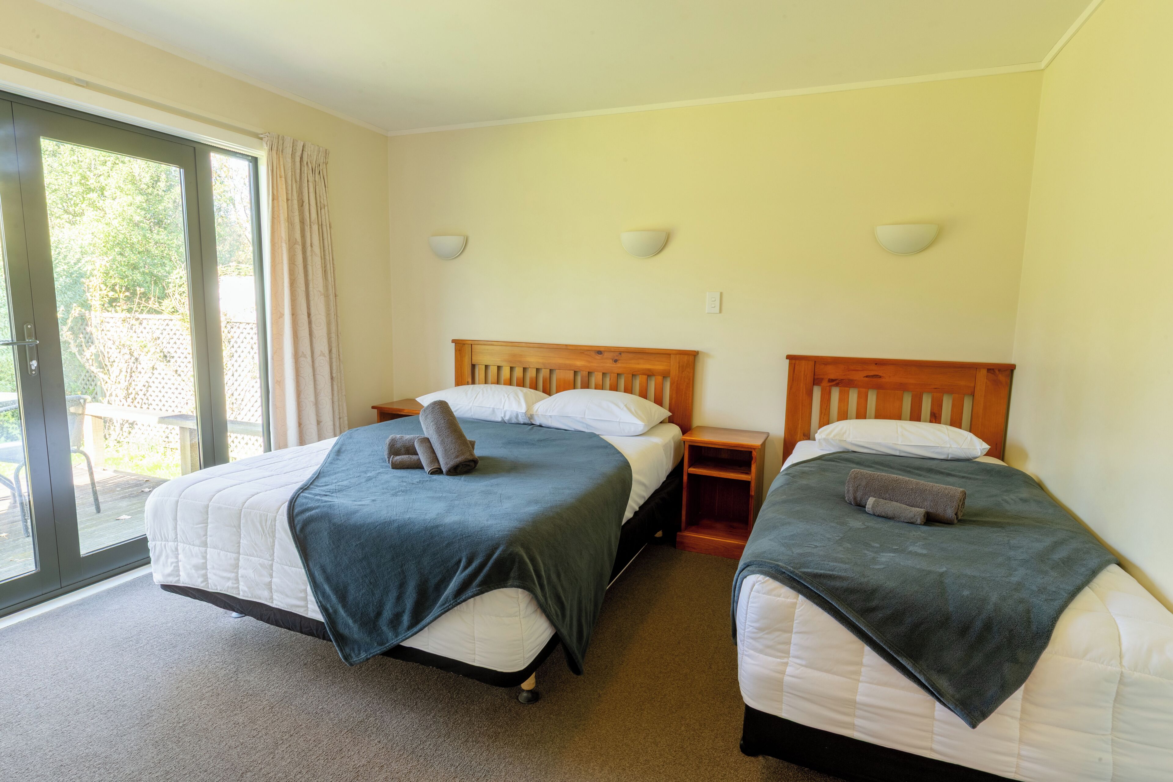 Une chambre d’hôtel avec deux lits, une porte-fenêtre coulissante donnant sur l’extérieur et une petite table de chevet en bois placée entre les lits.
