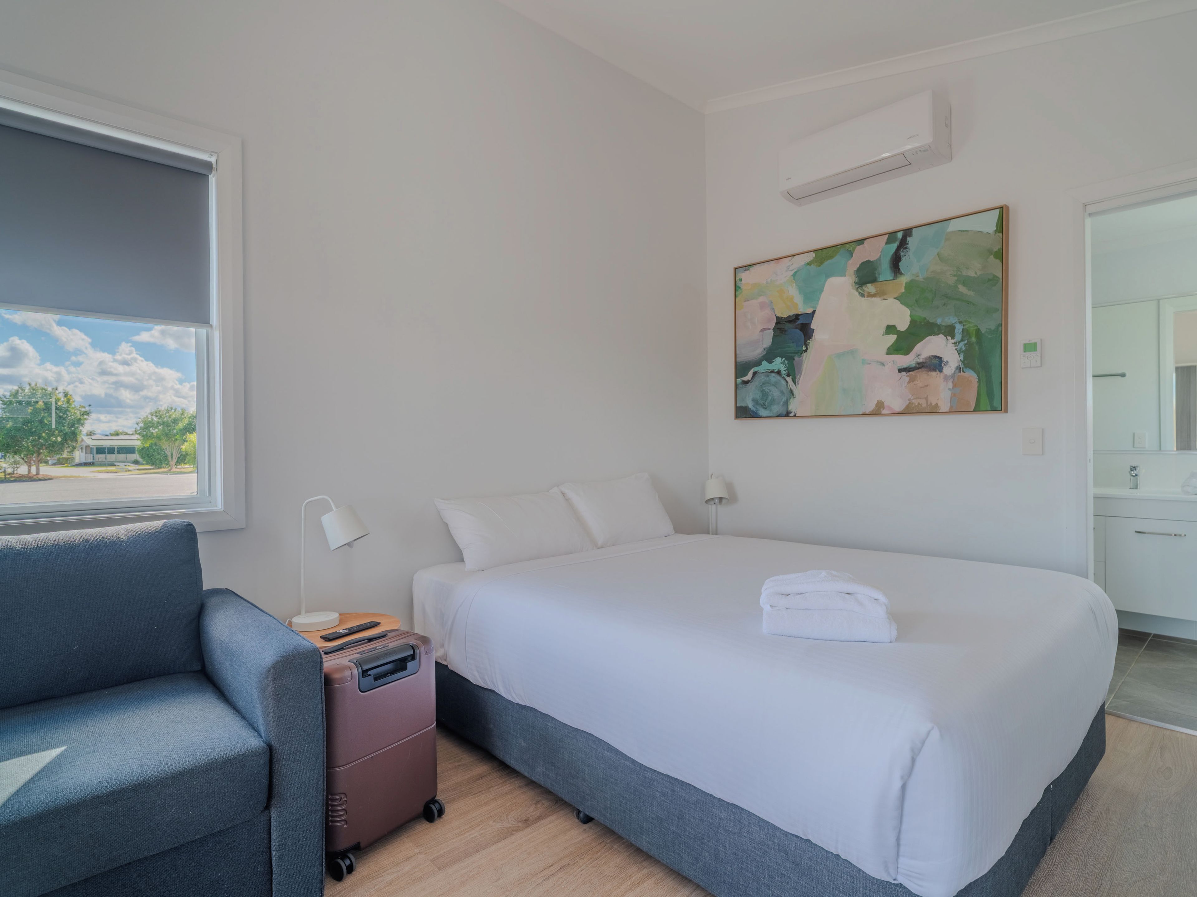 A hotel room with a bed, a desk, a chair, a suitcase, and a window with a view of the outside.
