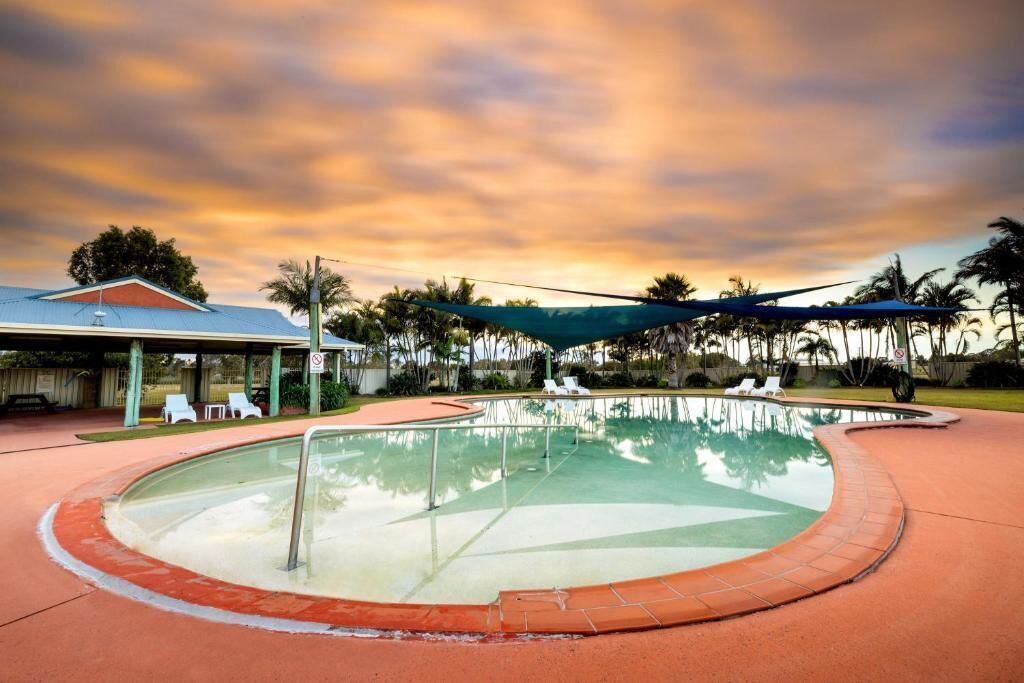 A swimming pool with a curved edge, surrounded by a red deck area. There are lounge chairs and a covered seating area in the background.