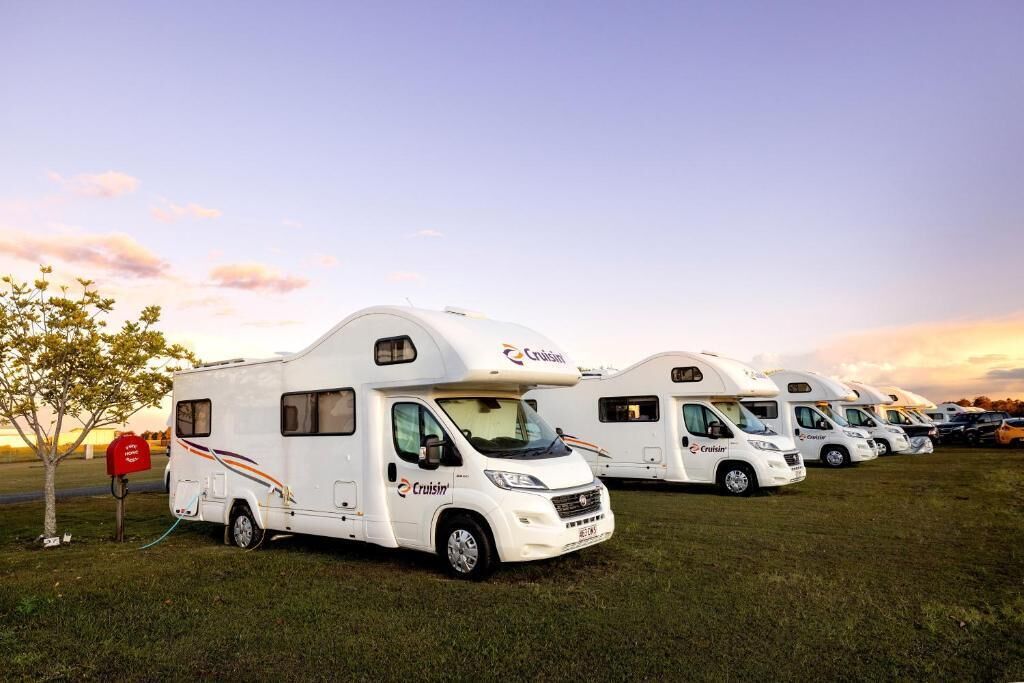 A row of white motorhomes parked in a grassy field with a sunset in the background.