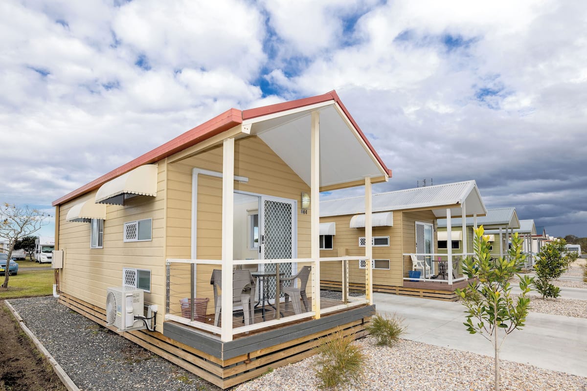 Una casa móvil de un solo nivel con porche cubierto, una pequeña terraza y una unidad de aire acondicionado visible.