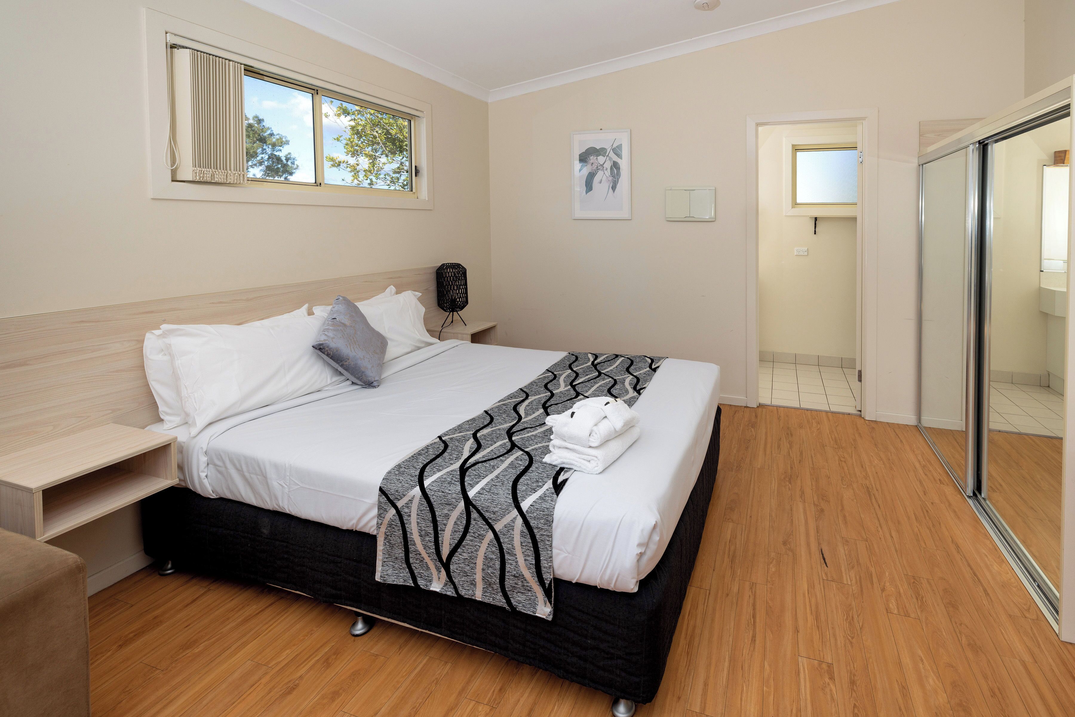 Un letto rifatto con cura, una coperta e dei cuscini decorati, un comodino con una lampada e una finestra con tende.