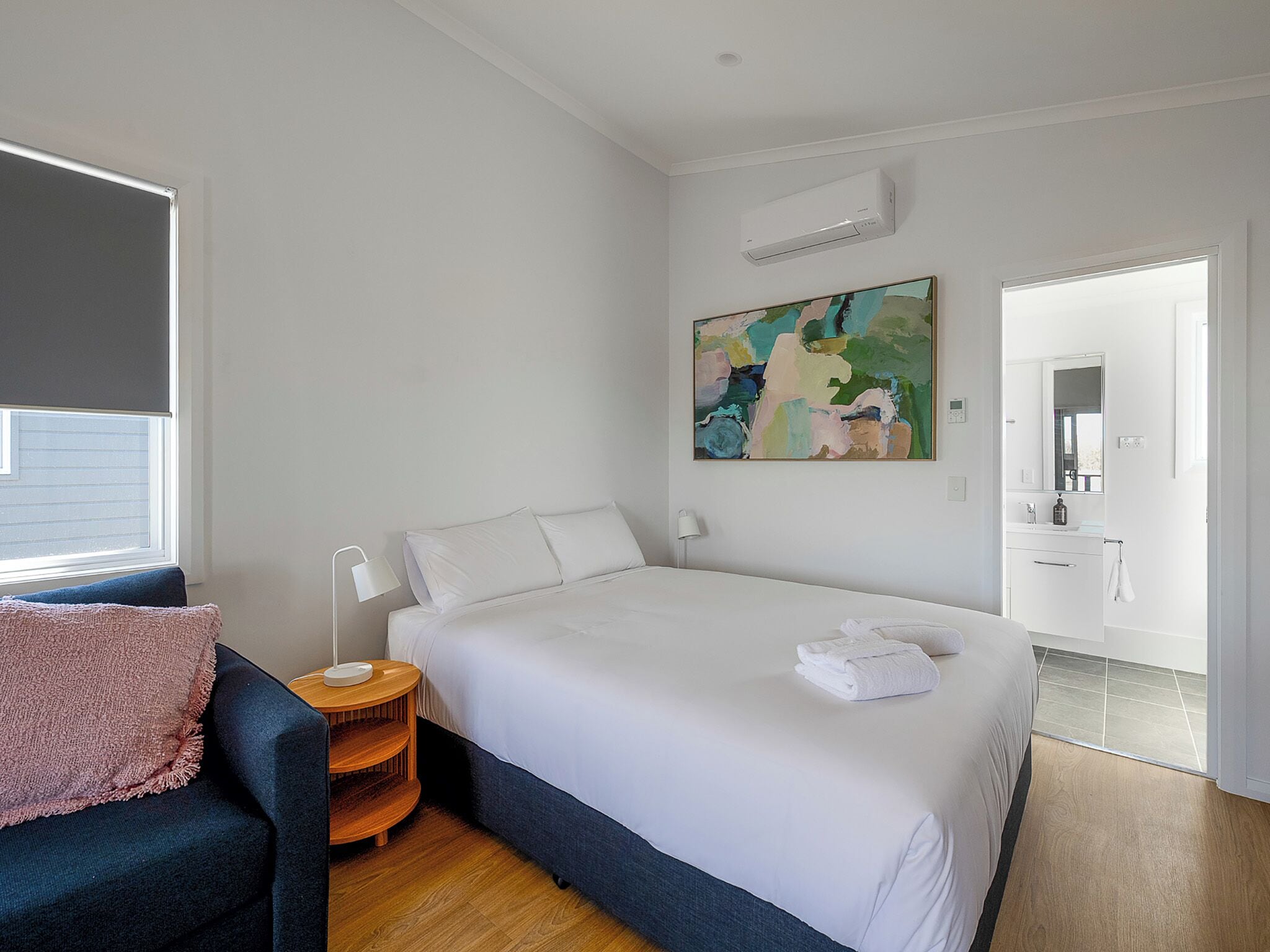 A modern hotel room with a large bed, a desk, a chair, a painting, and a bathroom visible through an open door.