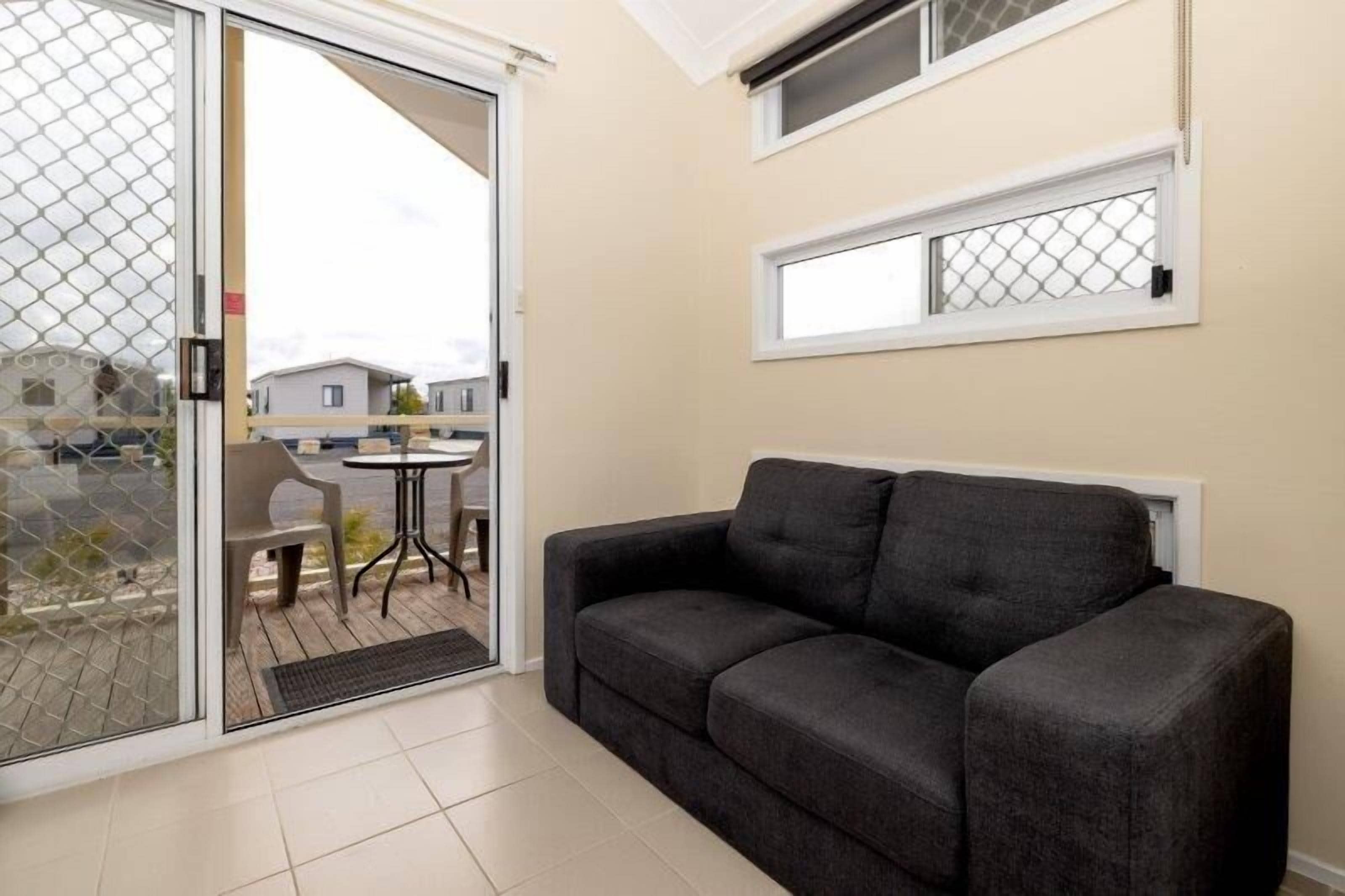 A room with a sliding glass door leading to an outdoor area, a grey sofa, a small round table, and a window with a white frame.