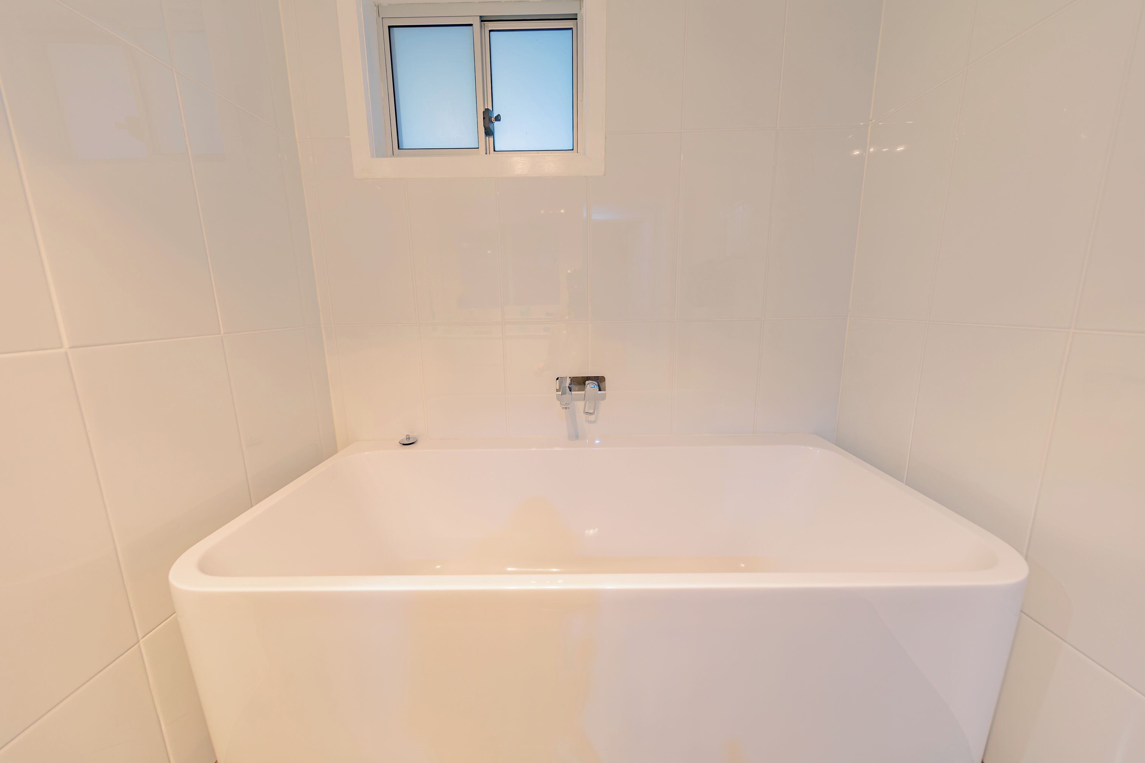 A bathroom with a white bathtub, a window above it, and white tiled walls.