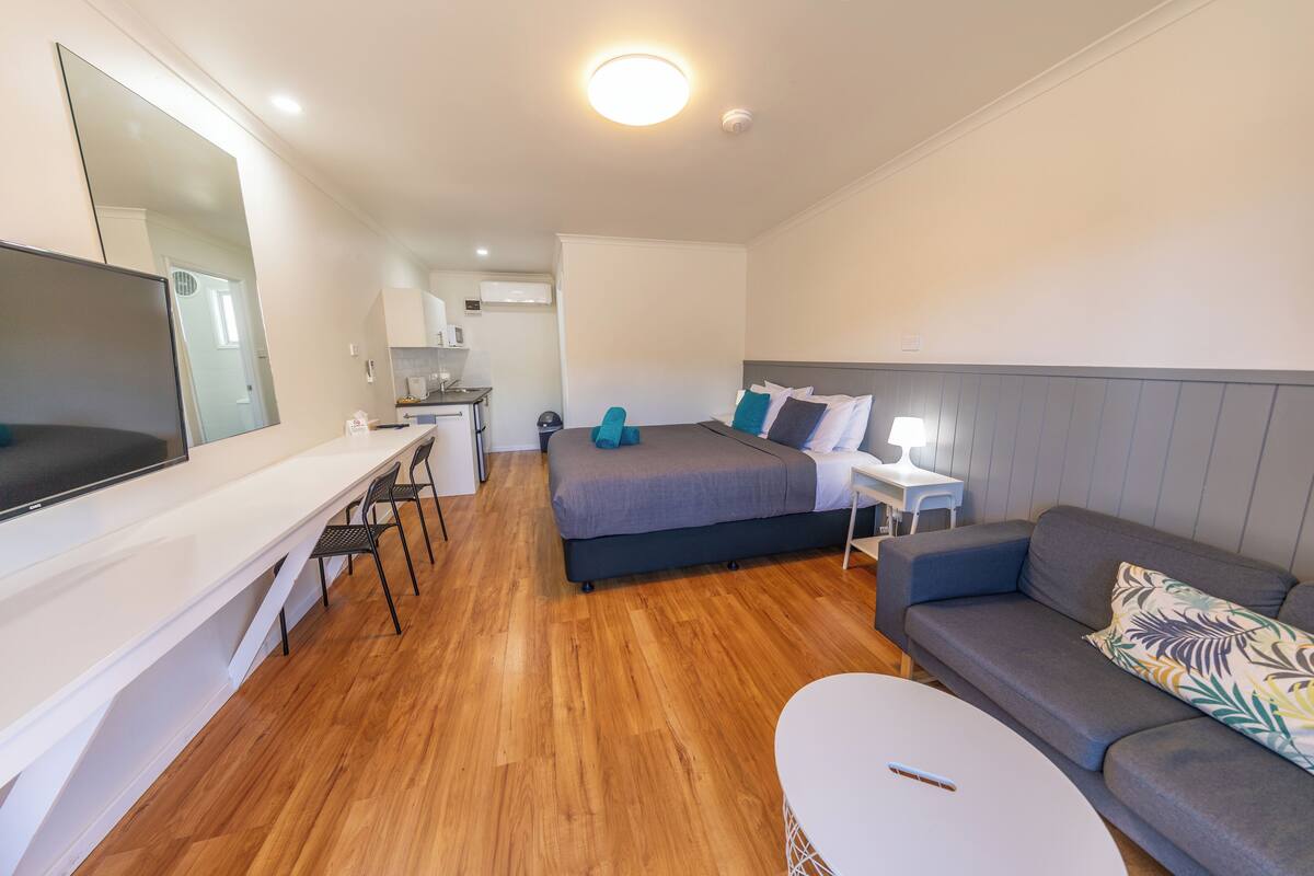 A modern hotel room with a bed, a sofa, a desk with a chair, a TV, and a small kitchen area.