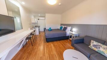 A modern hotel room with a bed, a sofa, a desk with a chair, a TV, and a small kitchen area.