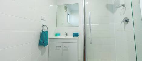 Un baño moderno con azulejos blancos, mampara de ducha de vidrio, lavamanos con espejo y toallero.