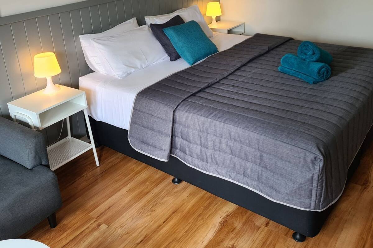 A neatly made bed with pillows, a bedside lamp, a rolled towel, and a picture on the wall.