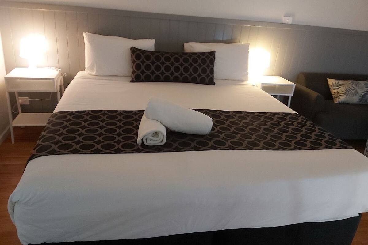 A neatly made bed with a patterned bedspread, white pillows, and a rolled towel on the bed. There are two bedside tables with lamps, a chair with a cushion, and a wall-mounted light fixture.