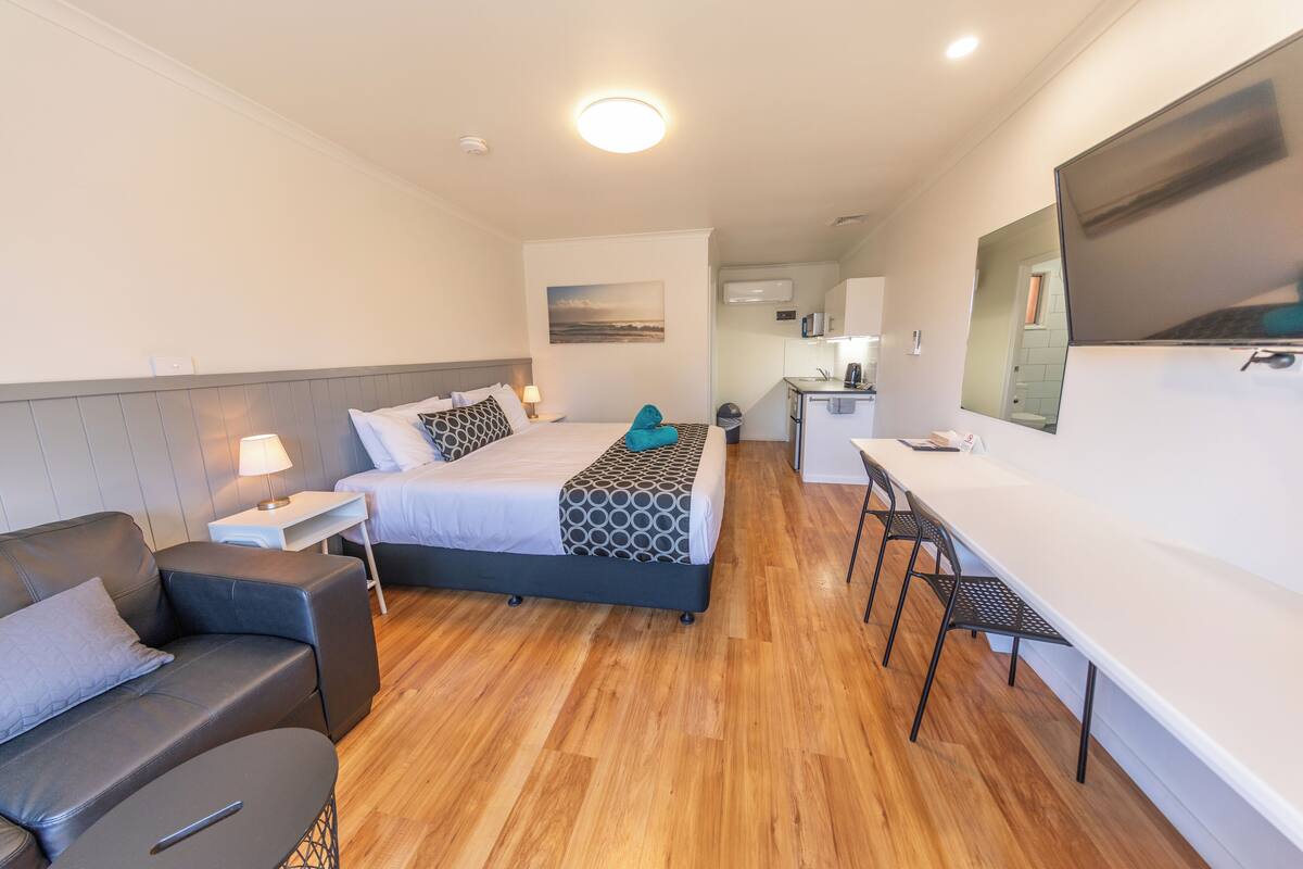 A modern hotel room with a bed, a desk, a chair, and a small kitchenette.