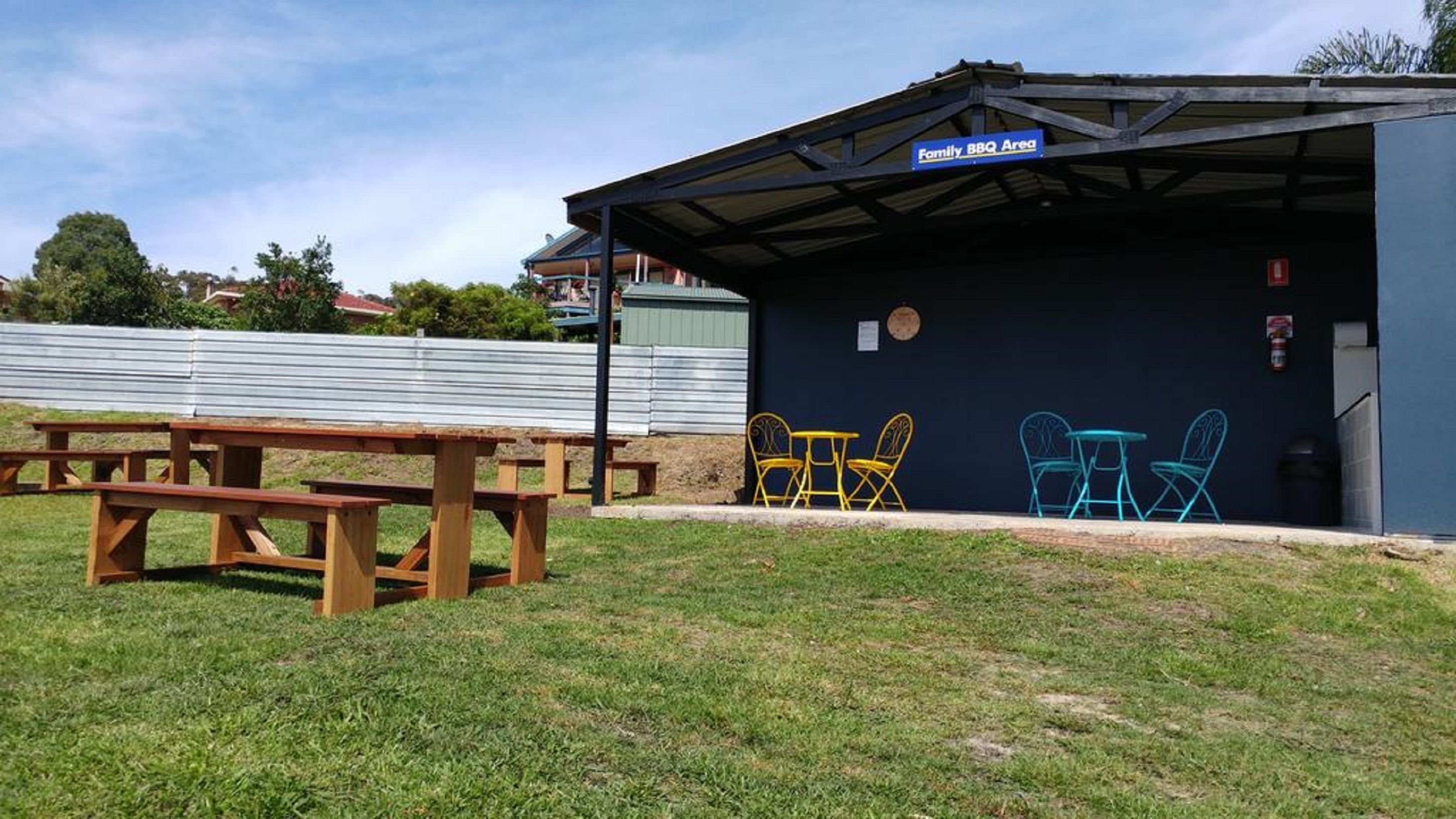 Un espacio al aire libre techado con una mesa de picnic y sillas de colores, señalizado como Área de Barbacoa Familiar.