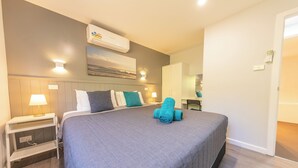 Una cama bien hecha con colcha gris y toallas azules, una mesita de noche con lámpara, aire acondicionado de pared y un cuadro de un paisaje.