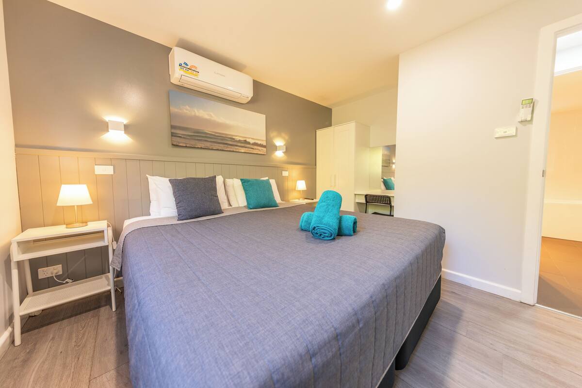 A neatly made bed with a grey bedspread and blue towels, a nightstand with a lamp, a wall-mounted air conditioner, and a painting of a landscape.