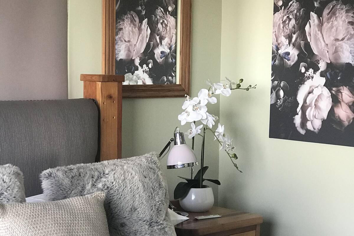 Une chambre à coucher avec un lit, des oreillers, une table de chevet avec une lampe, un miroir et des œuvres d’art florales accrochées au mur.