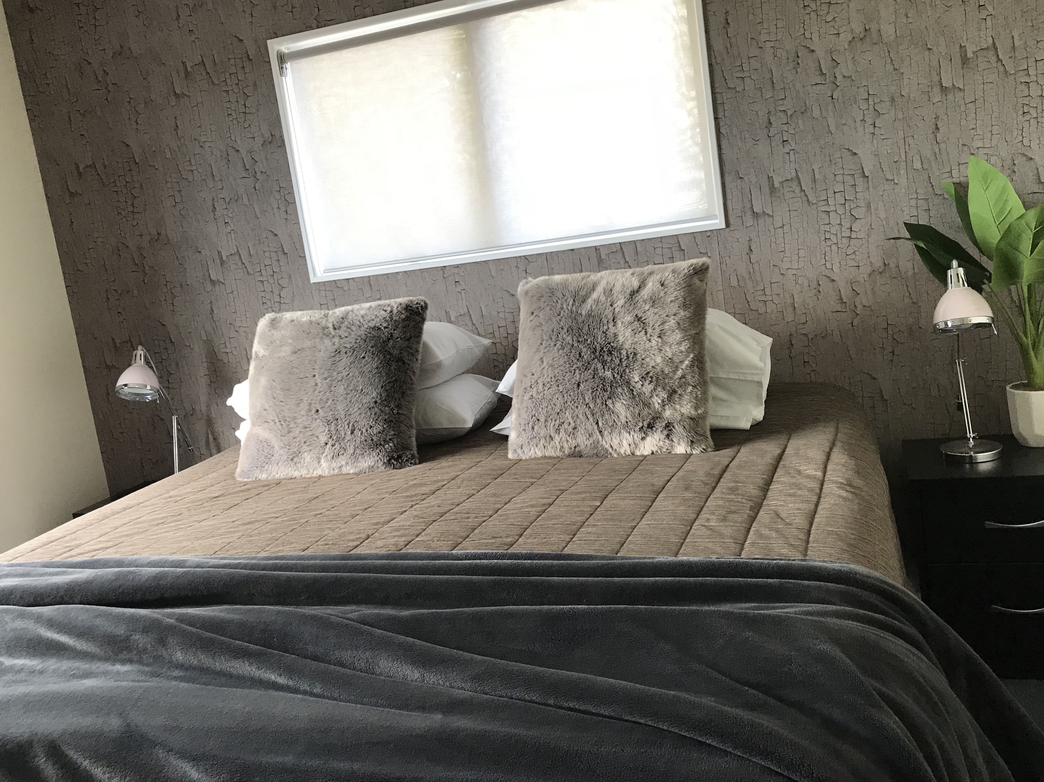 Una cama bien tendida con una manta gris, dos almohadas grises y una blanca. Hay una mesita de noche con una lámpara y una planta en maceta.