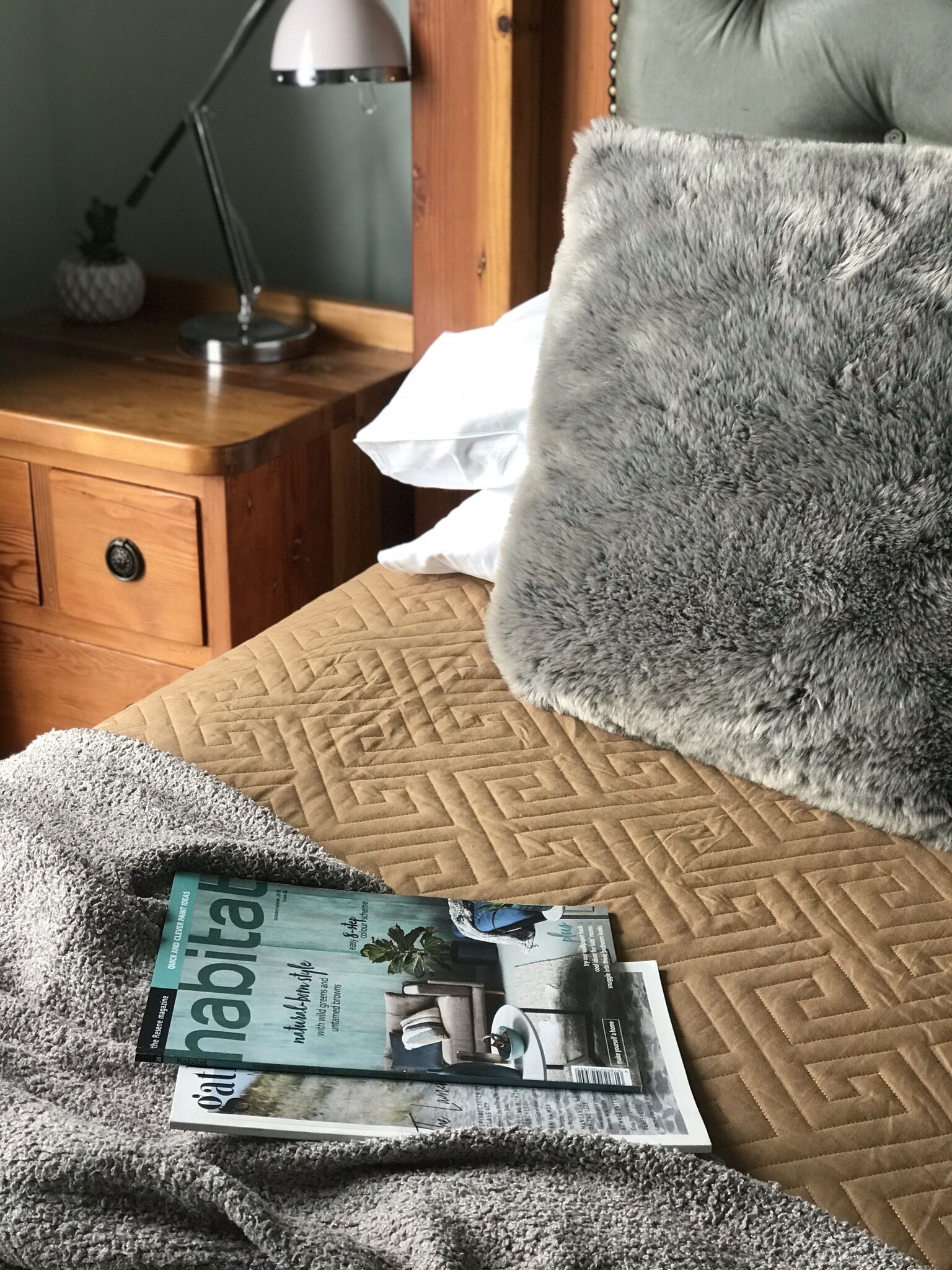 A neatly made bed with a quilted headboard, a fluffy pillow, and a magazine titled "habitat".