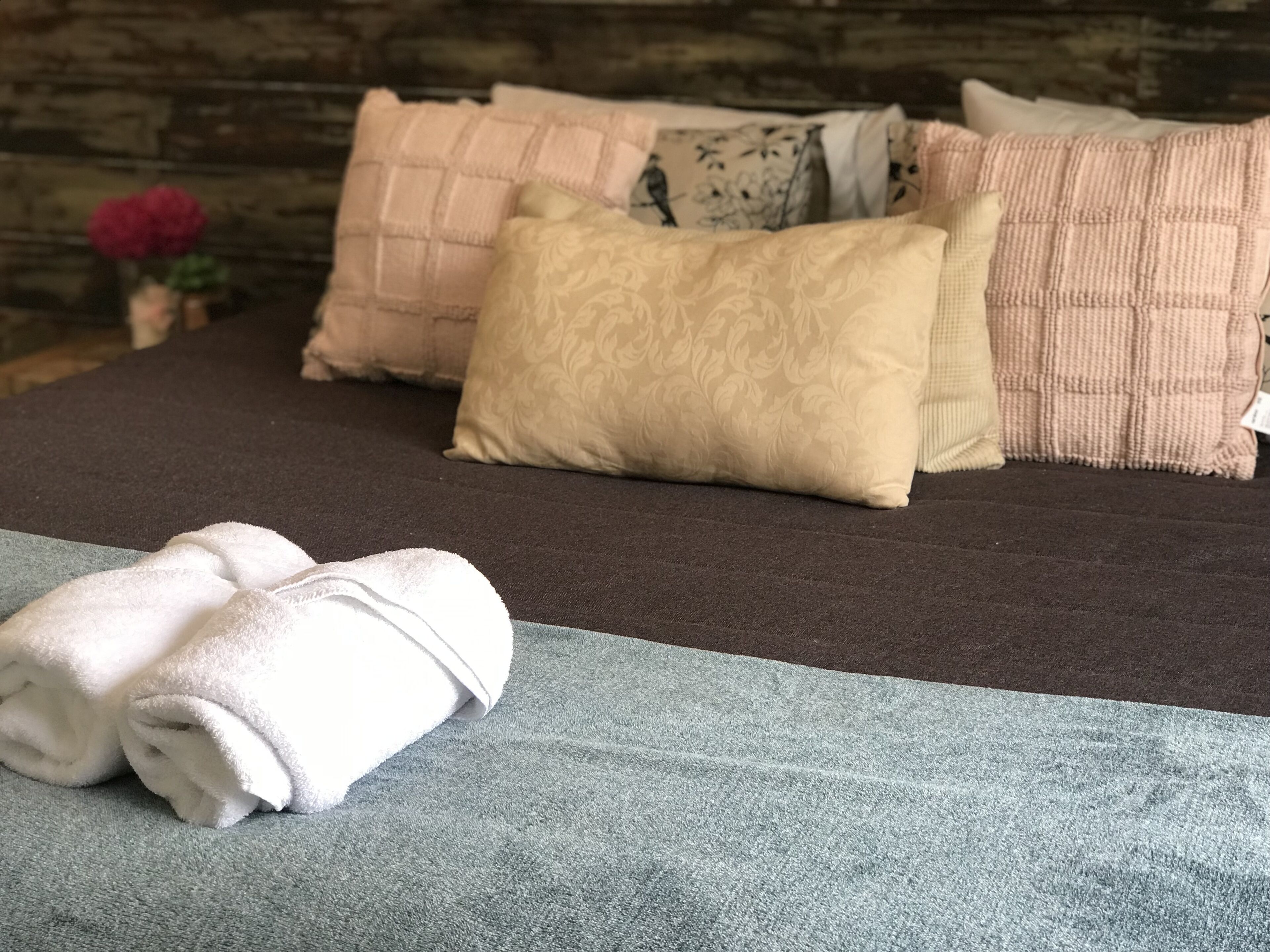 Una cama bien hecha con una colcha gris, una toalla blanca doblada y varios cojines decorativos de diferentes estampados y colores.