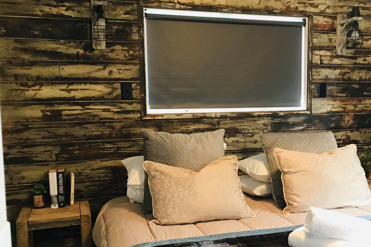 Une chambre avec un mur en bois, un lit avec une literie beige, une table de chevet avec des livres et un téléviseur fixé au mur.