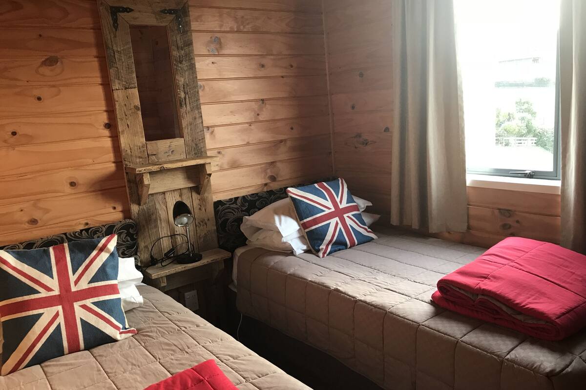 Une chambre confortable, aménagée dans le style d’un chalet, avec deux lits, un mur en bois, une fenêtre avec des rideaux et une petite table avec une lampe.