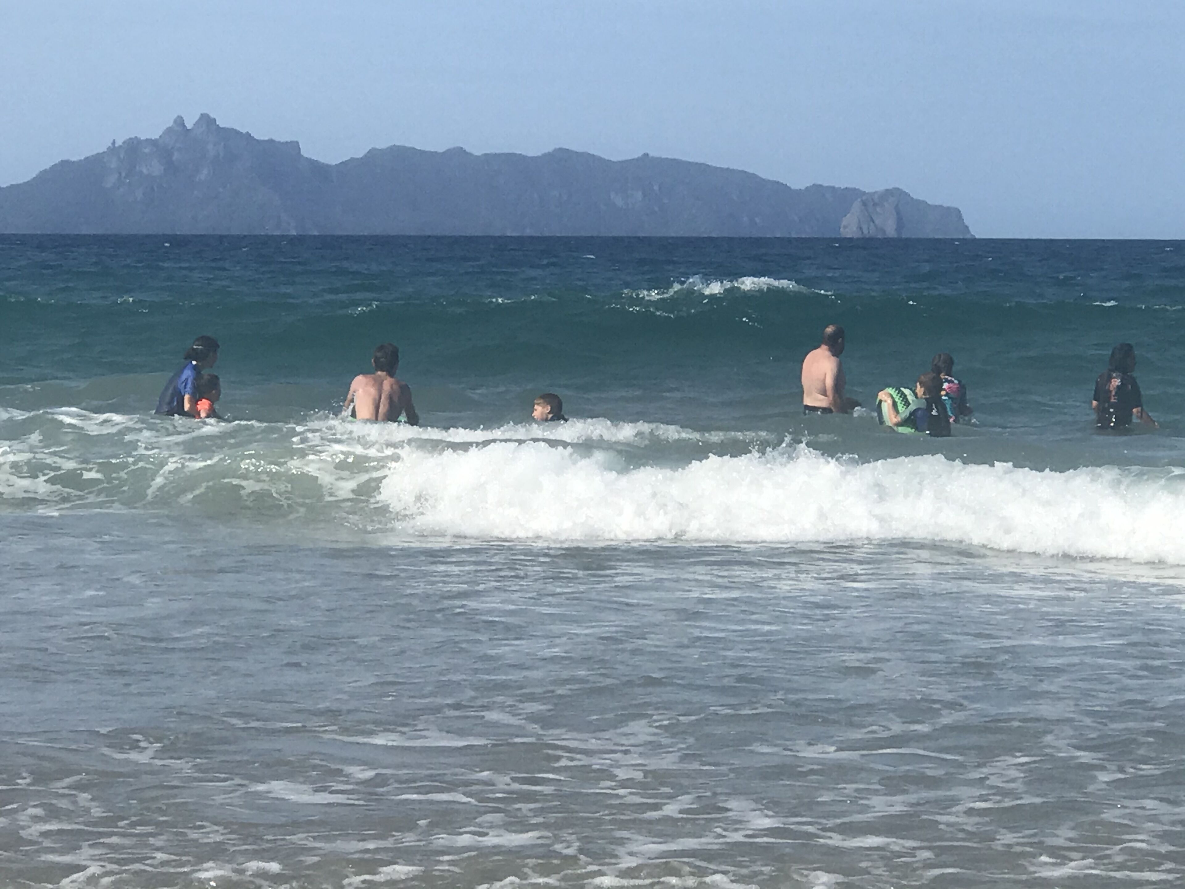 A beach scene with people in the water, some with surfboards, and mountains in the background.