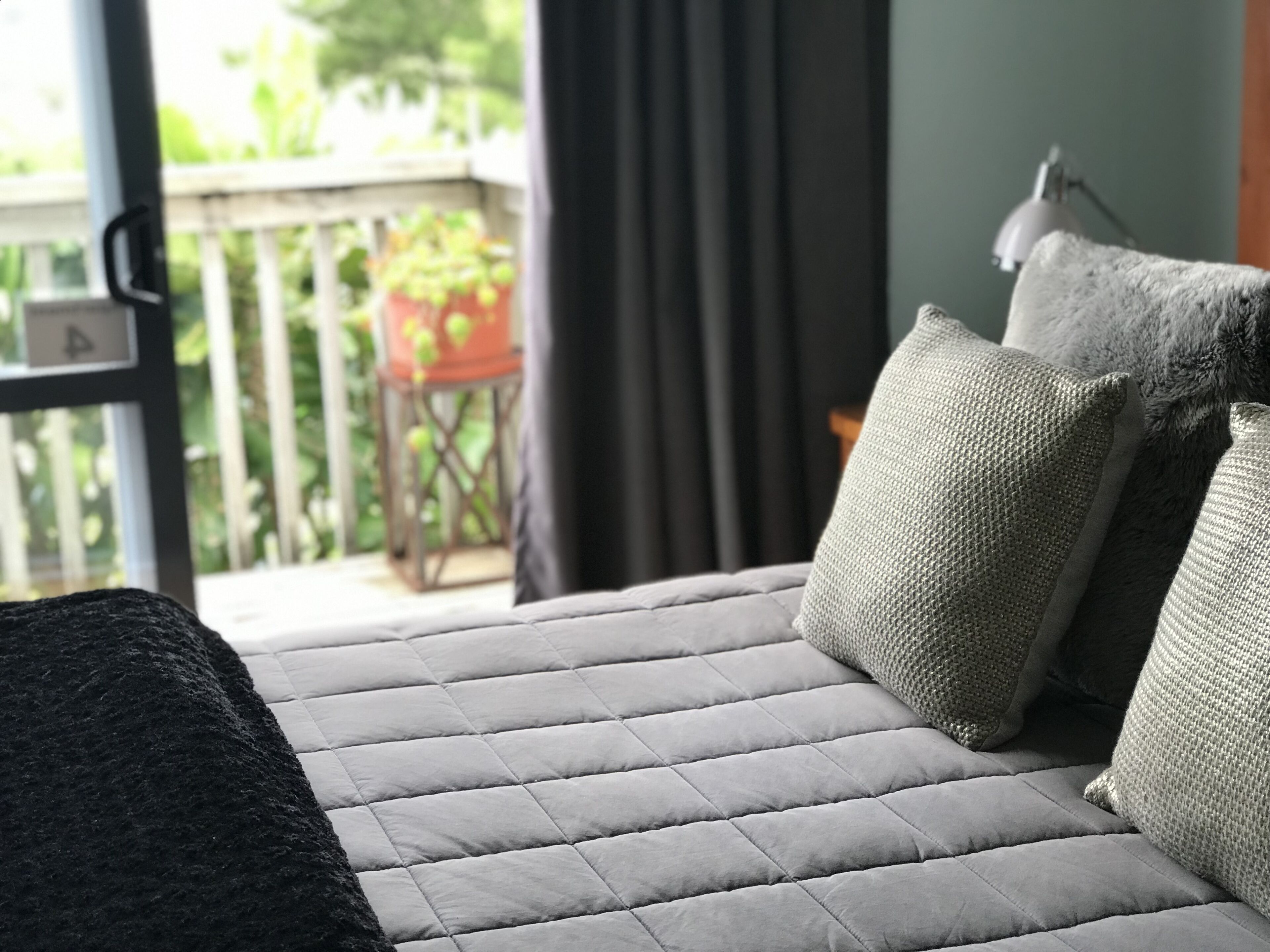 A bed with a grey quilted headboard and pillows, a black throw blanket, and a view of a balcony with a chair and plants.