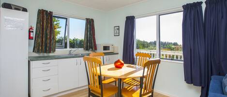 A modern kitchen with white cabinets, a wooden dining table, and chairs. There is a window with curtains, a microwave, and a fire extinguisher.