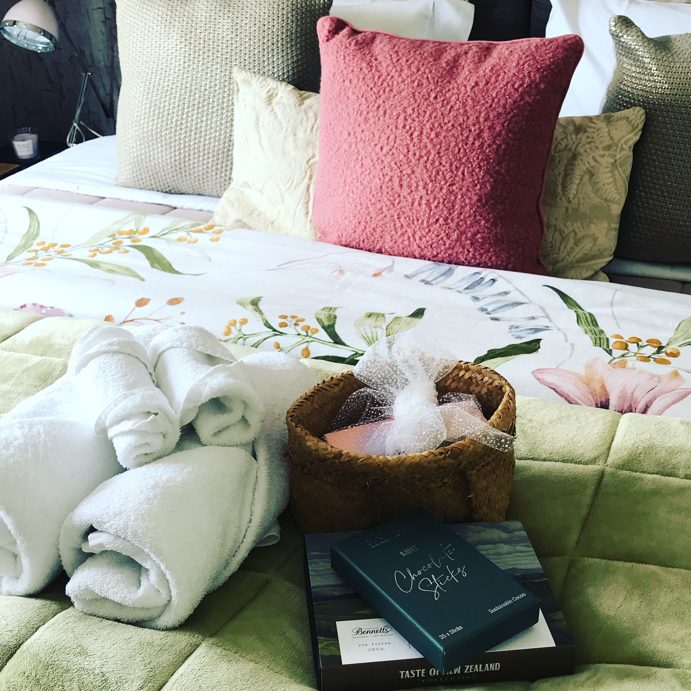 A neatly made bed with a floral bedspread, rolled white towels, a basket of toiletries, and a book titled 'Mint Chocolate Sticks'.