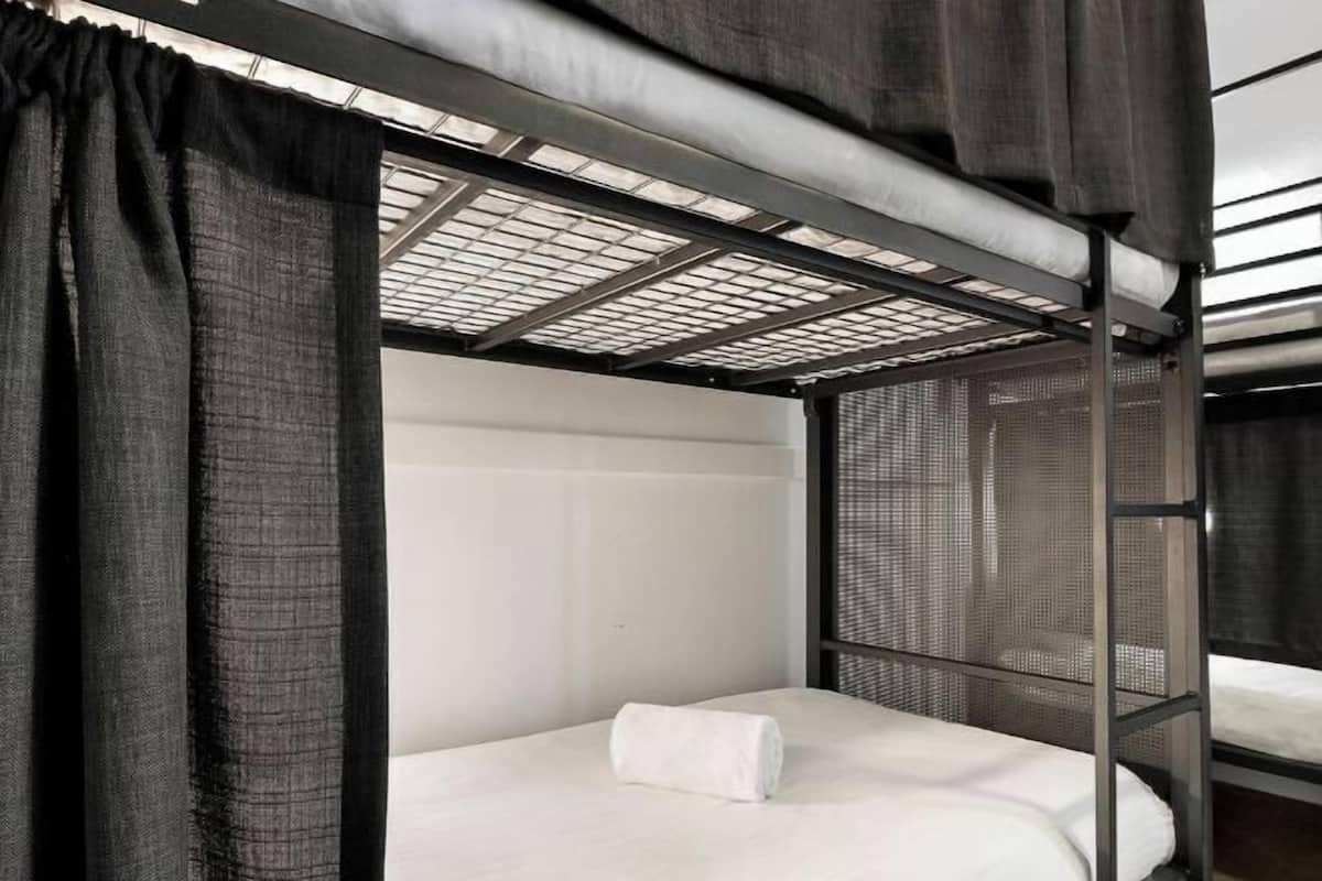 Una cama litera con colchón blanco y una toalla blanca enrollada, cabecero de malla metálica y ventana con vistas al cielo.