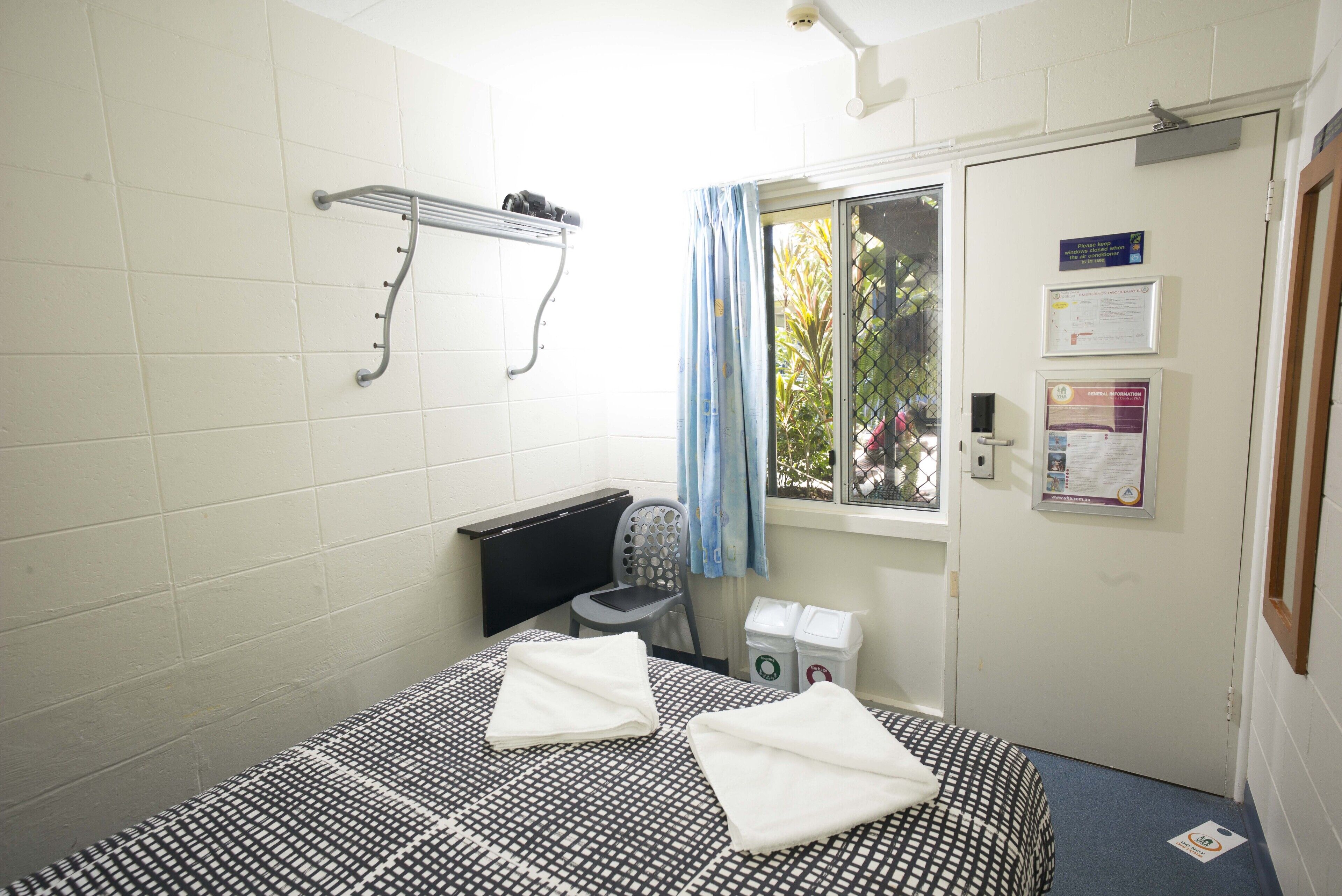 A small, clean room with a bed, a chair, a window with a curtain, a radiator, and a door with informational posters.