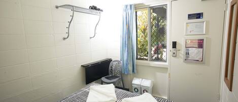 A small, clean room with a bed, a chair, a window with a curtain, a radiator, and a door with informational posters.