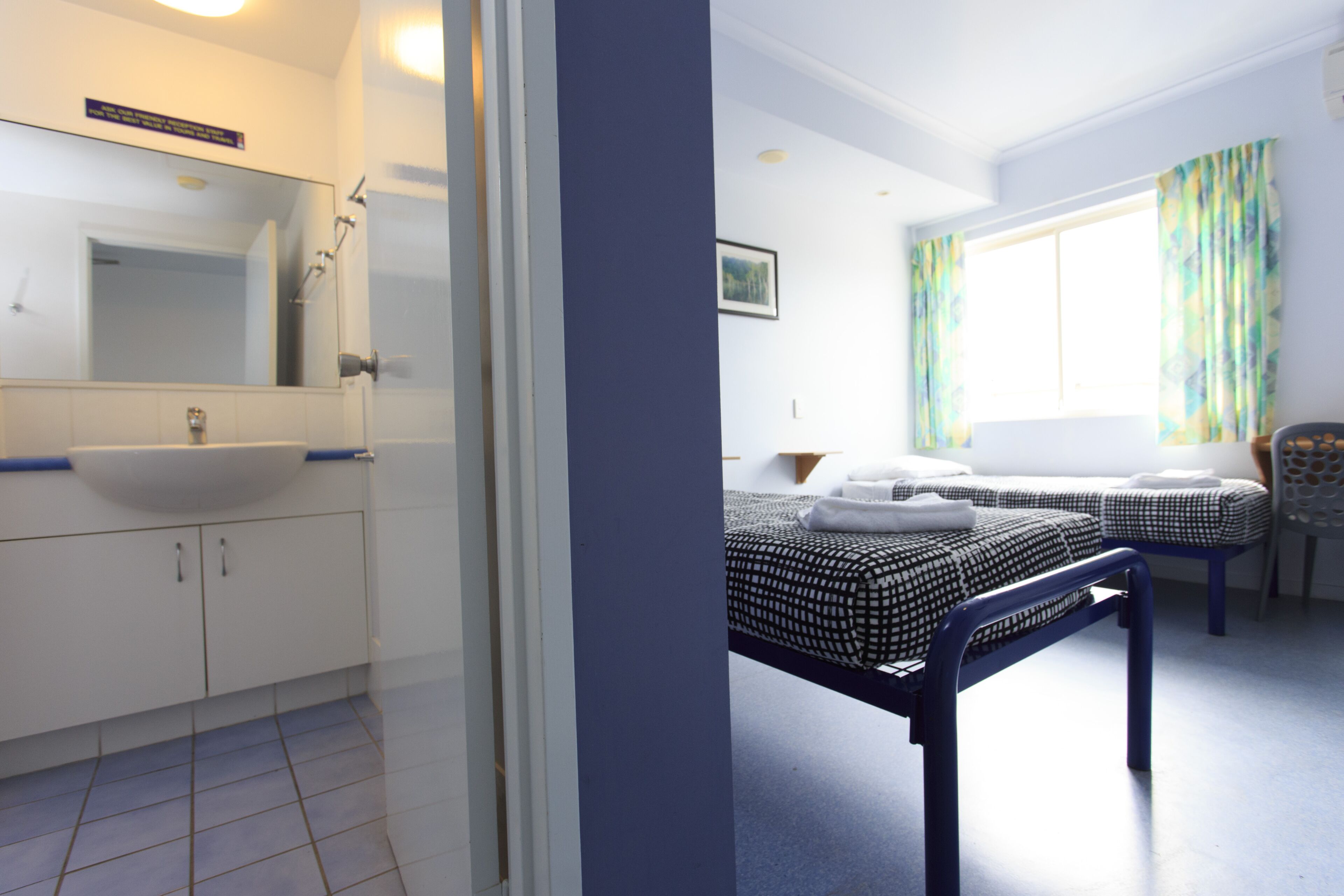 A hotel room with a bathroom, a bed with a checkered bedspread, a chair, and a window with curtains.