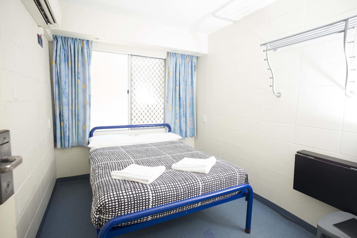 Una cama individual con una colcha estampada, estructura de metal azul, una ventana con cortinas azules, un radiador fijado a la pared y un cubo de basura pequeño.