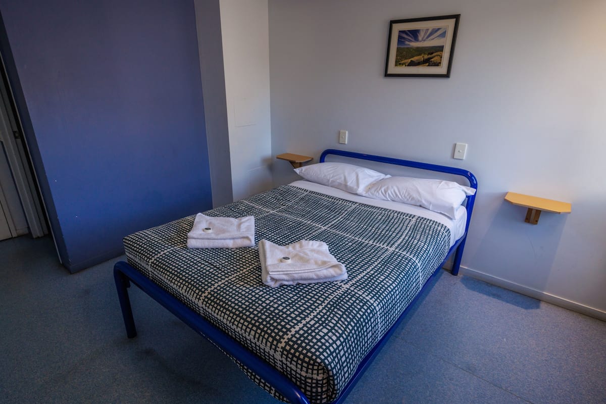 Una cama individual con una colcha a cuadros, dos almohadas y dos toallas dobladas. Hay una mesita de noche de madera a cada lado de la cama.