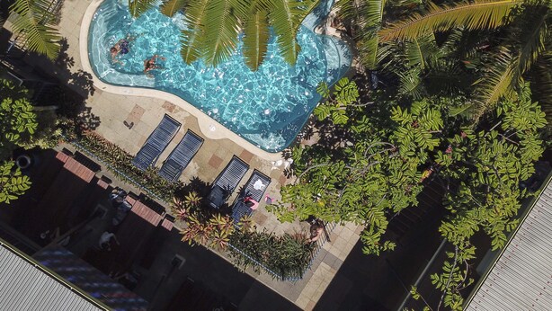 Vista aérea de una piscina rodeada de palmeras, con sillas de descanso y un área de terraza con baldosas.