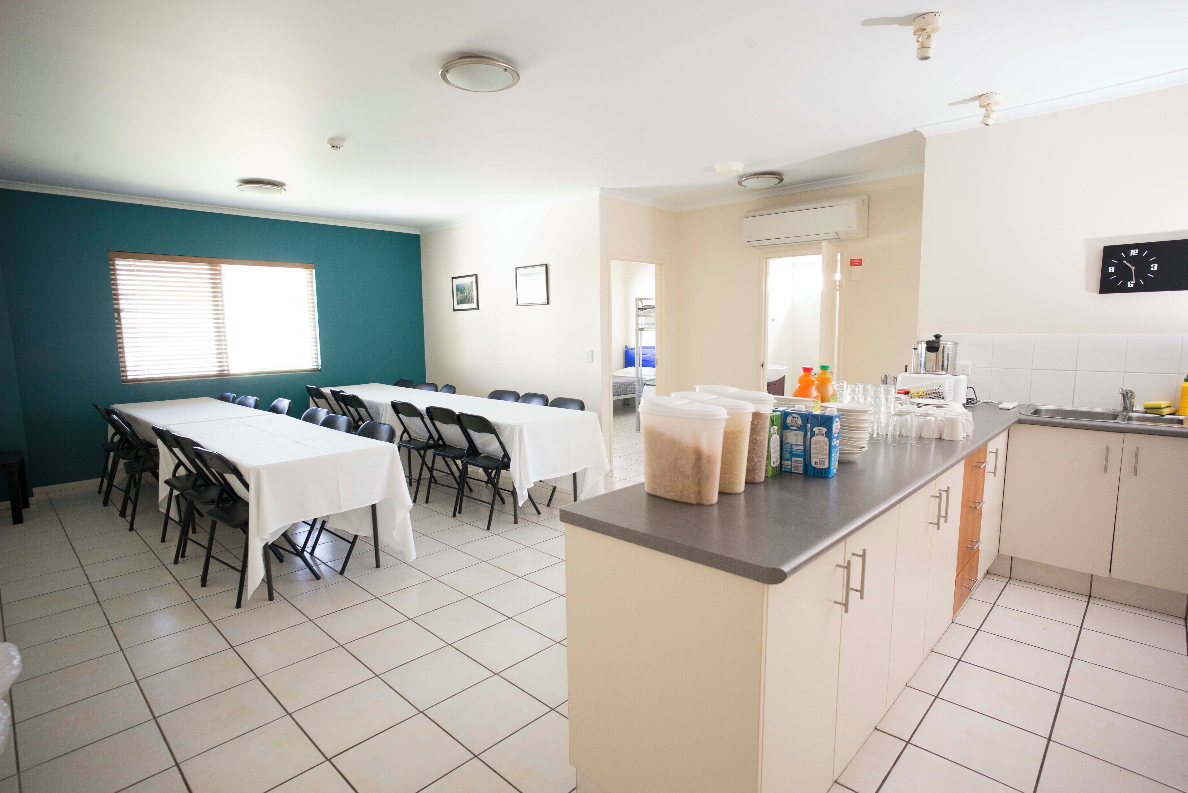 Un espace repas avec une longue table et des chaises, une cuisine avec un comptoir et des placards, et une fenêtre avec des stores.