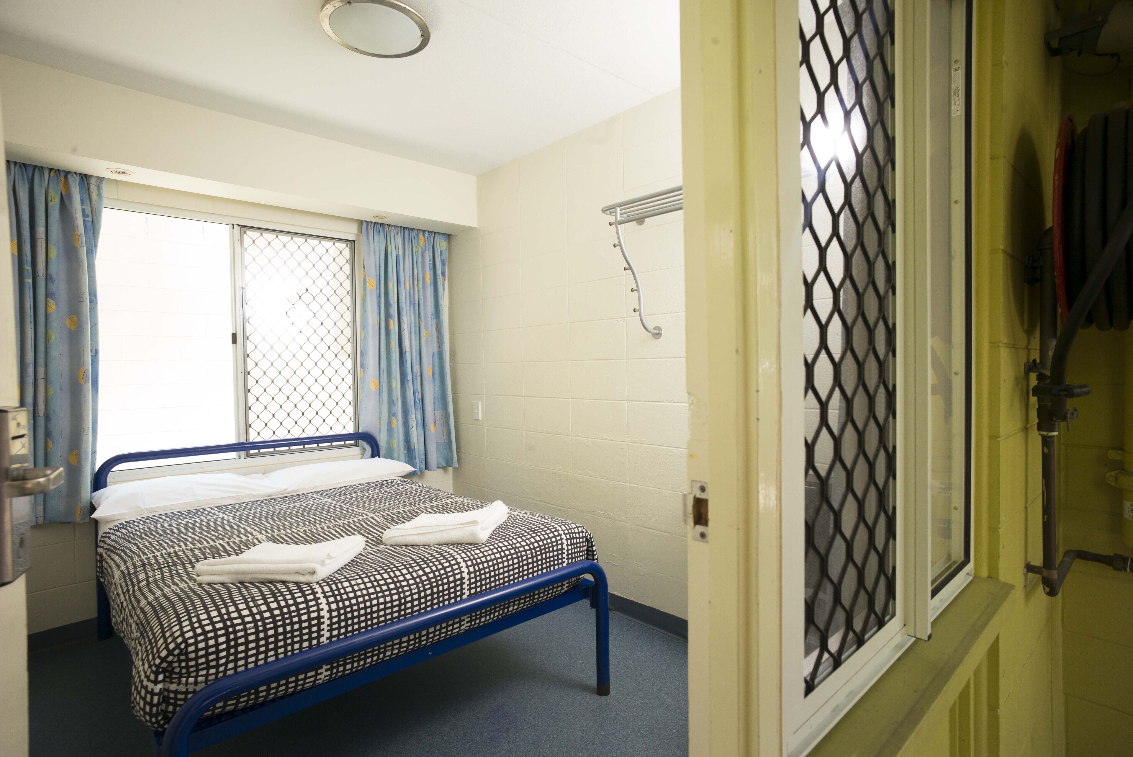 Een enkel bed met een geruit beddegoed en een opgevouwen handdoek, een raam met gordijnen, en een metalen airconditioning op de muur.