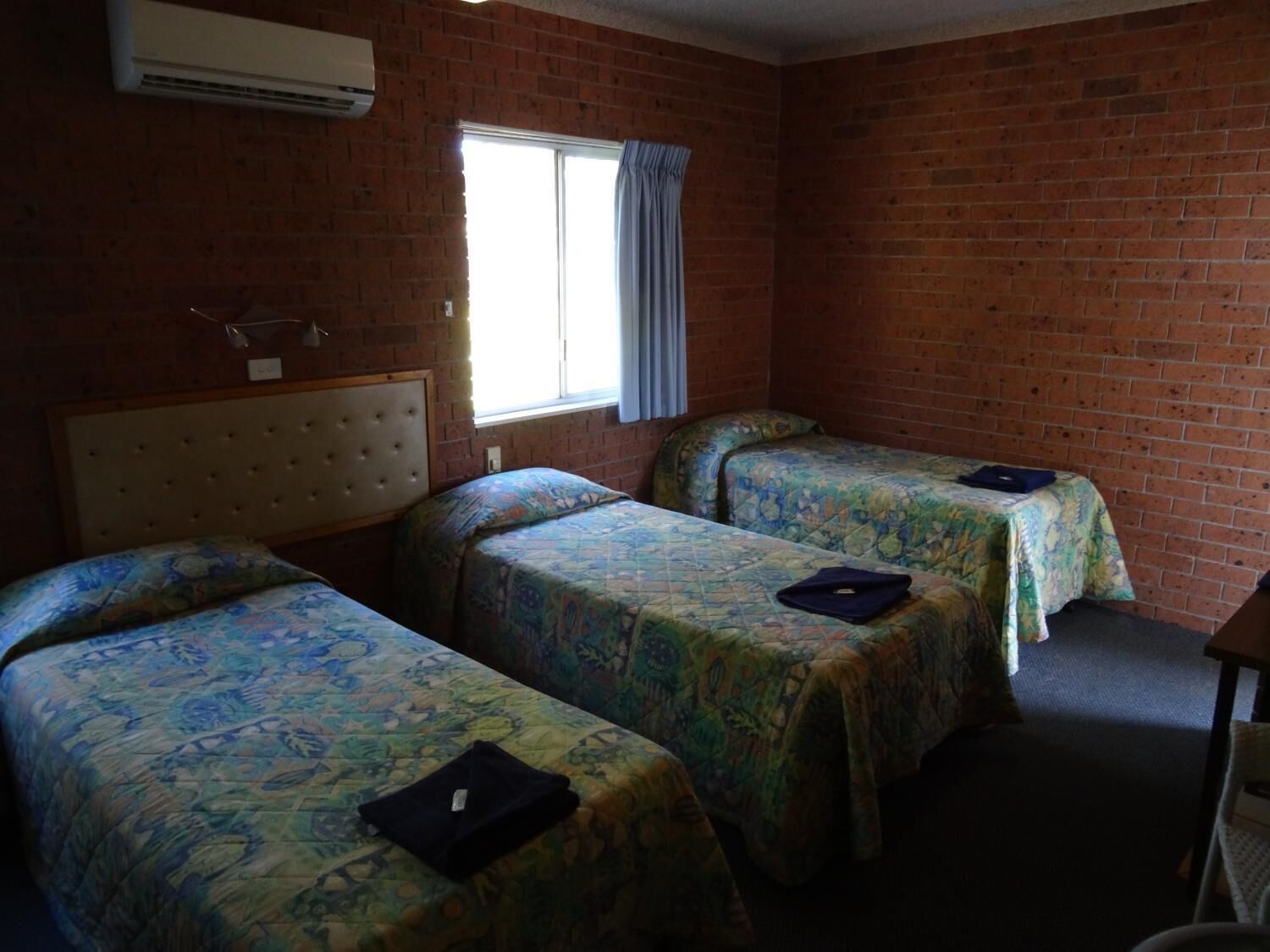 A room with two single beds, a headboard, a window with curtains, and an air conditioning unit.