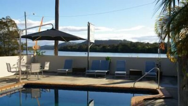 A swimming pool with lounge chairs and an umbrella, surrounded by a concrete deck and a view of a body of water and hills. - Chinderah Motel (Chinderah)